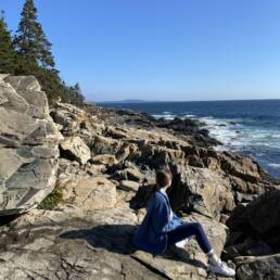 girl sitting on rocky beach overlooking water with pine trees in the distance